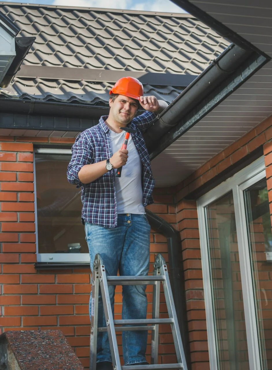 Smiling carpenter posing with hammer on step ladder under the roof